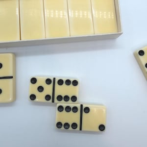 Puede incluir: Dominós de color marfil con puntos negros, algunos en una caja, otros dispersos. Los dominós tienen un diseño clásico con una línea negra que divide las dos mitades. Un juego tradicional.