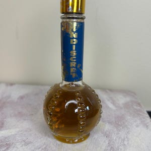 May include: A small, round, clear glass bottle with a gold cap and a blue label that reads "Indiscreet". The bottle is filled with a light brown liquid.