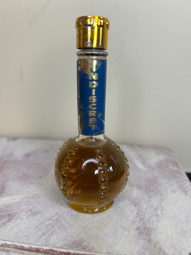 May include: A small, round, clear glass bottle with a gold cap and a blue label that reads "Indiscreet". The bottle is filled with a light brown liquid.