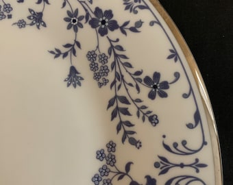 May include: A white ceramic plate with a blue and silver floral design around the edge. The plate has a gold rim.