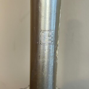 May include: Close-up of a silver bottle cap with a small, embossed, square logo. The logo is a stylized letter "L" with a small square inside.