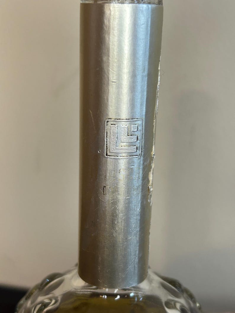 May include: Close-up of a silver bottle cap with a small, embossed, square logo. The logo is a stylized letter "L" with a small square inside.
