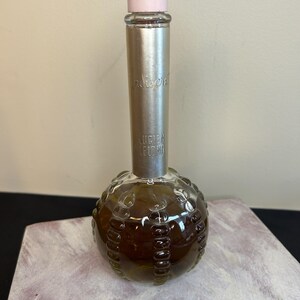May include: A vintage glass bottle of Indiscreet perfume by Helen Lebov. The bottle is round with a textured surface and a silver cap with a pink top. The bottle is filled with a brown liquid.