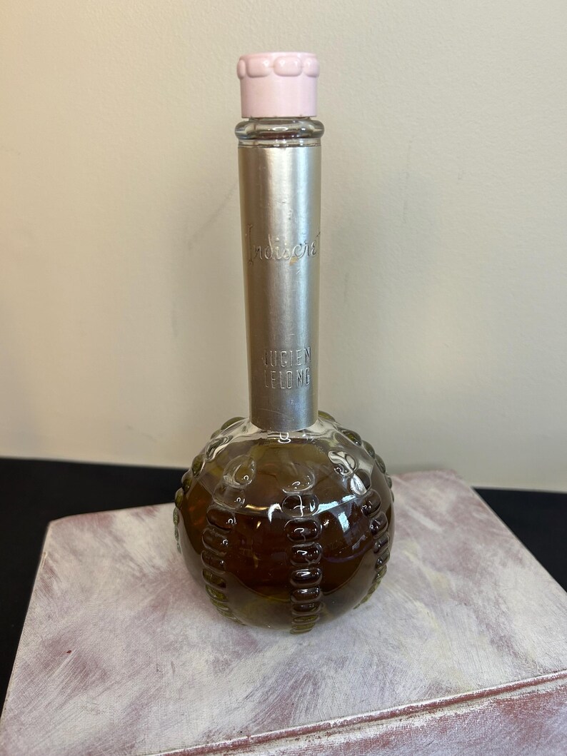 May include: A vintage glass bottle of Indiscreet perfume by Helen Lebov. The bottle is round with a textured surface and a silver cap with a pink top. The bottle is filled with a brown liquid.