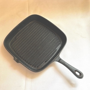 May include: A square, black cast iron grill pan with a ridged cooking surface. The pan has a long handle with a hole at the end for hanging. The pan is designed for grilling food and has a classic, rustic appearance.