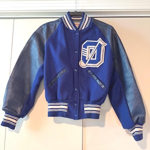 May include: A blue and navy varsity jacket with a white and navy striped collar, cuffs, and hem. The jacket features navy leather sleeves and a large white and navy letter "D" patch on the chest.