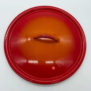 May include: A round, red and orange enameled pot lid with a small handle. The lid has a gradient color scheme, transitioning from a deep red at the edges to a bright orange in the center. The handle is a matching red.