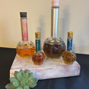 May include: Four vintage perfume bottles with round, textured glass bases. The bottles are filled with clear liquid and have different colored labels. The labels read "Balladine", "Signet", "Intrigue", and "Jamais" with a pink cap. The bottles are arranged on a white wooden surface.