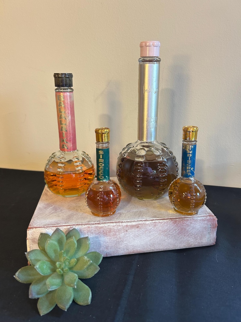 May include: Four vintage perfume bottles with round, textured glass bases. The bottles are filled with clear liquid and have different colored labels. The labels read "Balladine", "Signet", "Intrigue", and "Jamais" with a pink cap. The bottles are arranged on a white wooden surface.