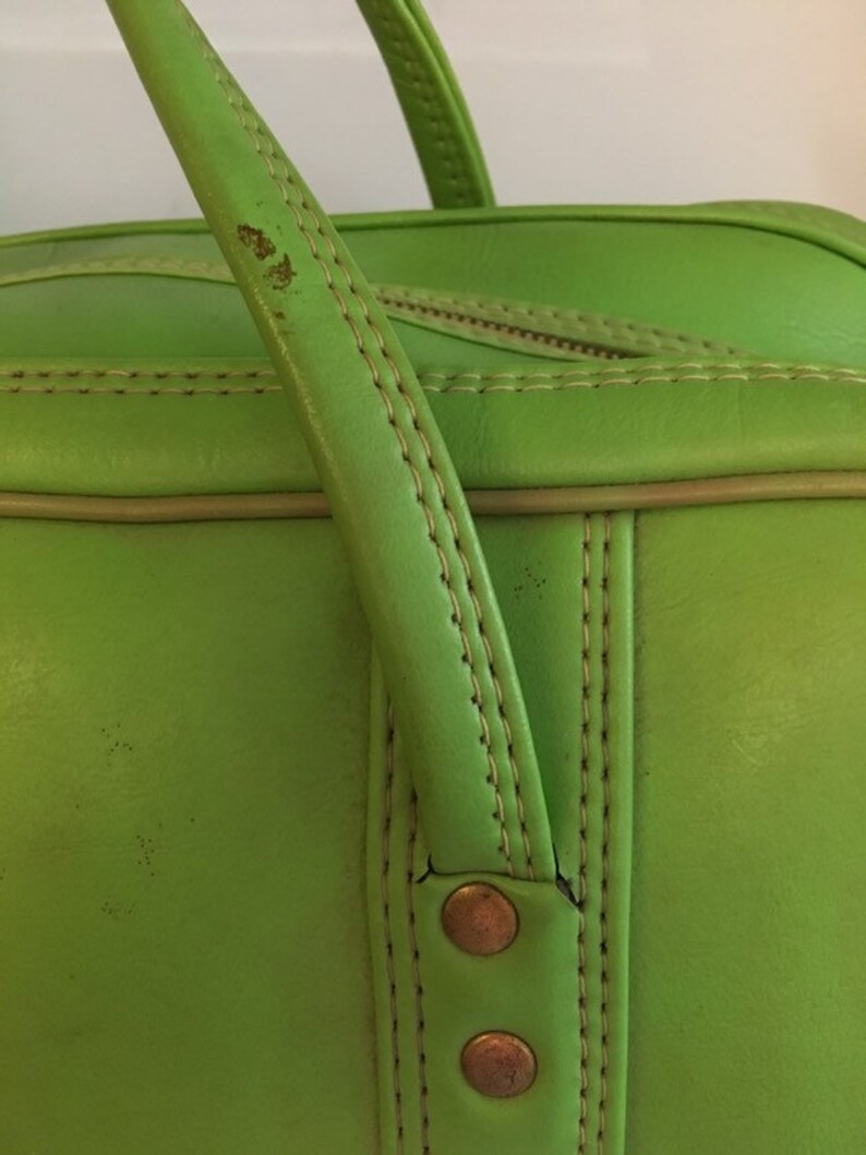 May include: Close-up of a bright green leather travel bag with a top handle and two brass rivets. The bag has a light brown trim and stitching.
