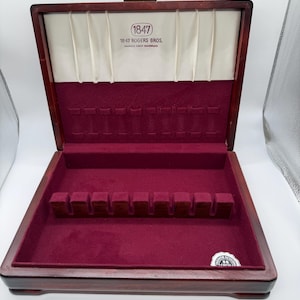 May include: Open wooden silverware chest with a burgundy velvet interior. The chest has a gold-colored clasp and the text "1847 Rogers Bros." is visible on the inside of the lid. The chest is designed to hold silverware.