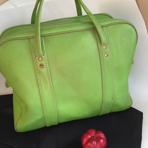 May include: A bright green leather travel bag with two top handles and gold-toned hardware. The bag is sitting on a black surface next to a red apple.