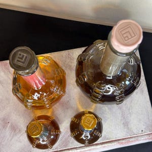 May include: Four glass bottles with textured surfaces and gold caps. The bottles are filled with a clear liquid and have a gold logo on the caps. The bottles are arranged on a white surface.
