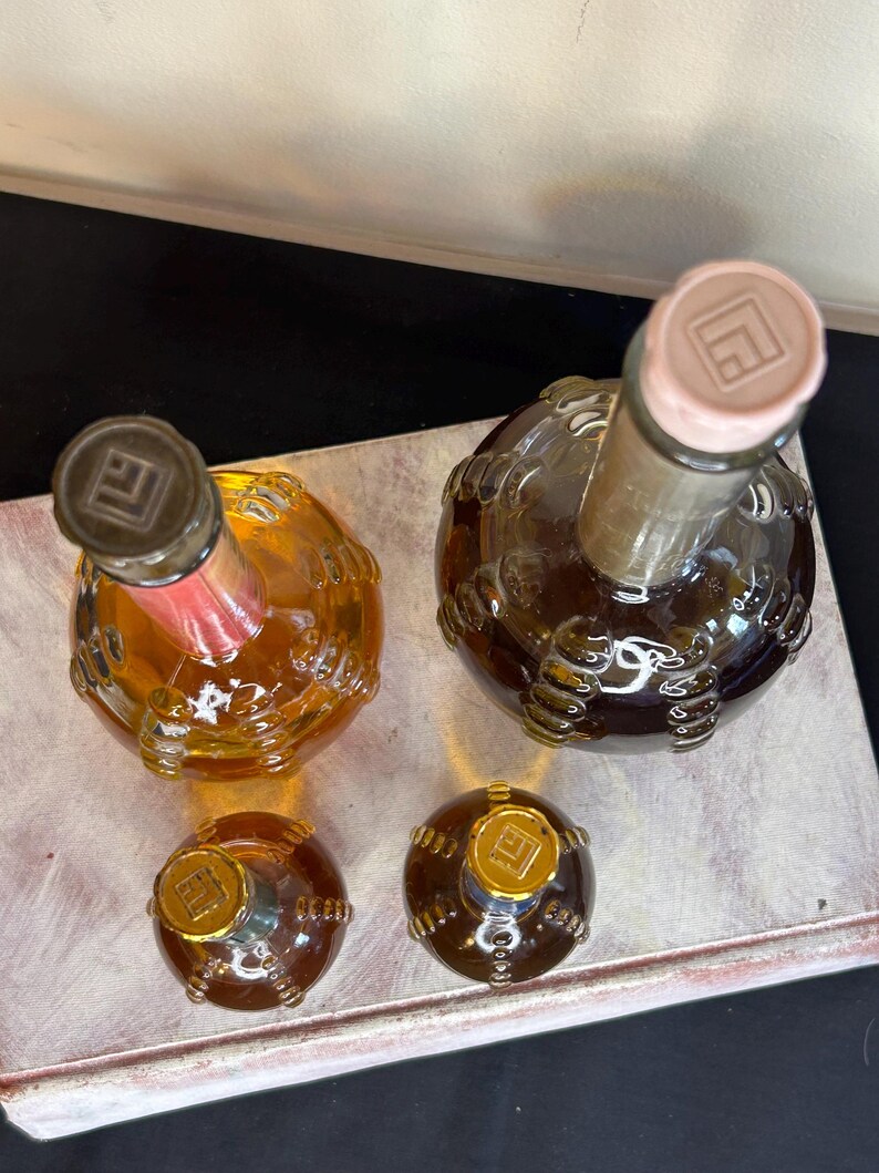 May include: Four glass bottles with textured surfaces and gold caps. The bottles are filled with a clear liquid and have a gold logo on the caps. The bottles are arranged on a white surface.