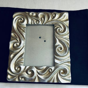 May include: A silver picture frame with an ornate design featuring swirls and scrolls. The frame is empty and ready for a photo.