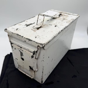 May include: A rectangular metal ammunition box painted white with visible signs of wear, including chipped paint revealing dark gray metal underneath. The box features a hinged lid with a central carrying handle and a side latch mechanism.