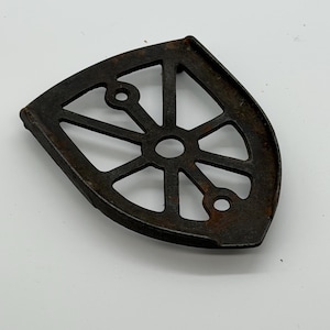 May include: A dark brown, cast iron trivet with a wheel-like design. The trivet is shaped like a quarter-circle with a series of spokes radiating from a central hole. Two smaller holes are located near the edge. The trivet is designed to protect surfaces from heat.