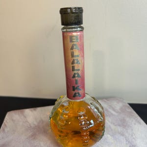 May include: A clear glass bottle with a textured, round base and a long, thin neck. The bottle contains a golden liquid and has a black cap. The bottle has a red label with the word "BALALAIKA" printed in gold letters.