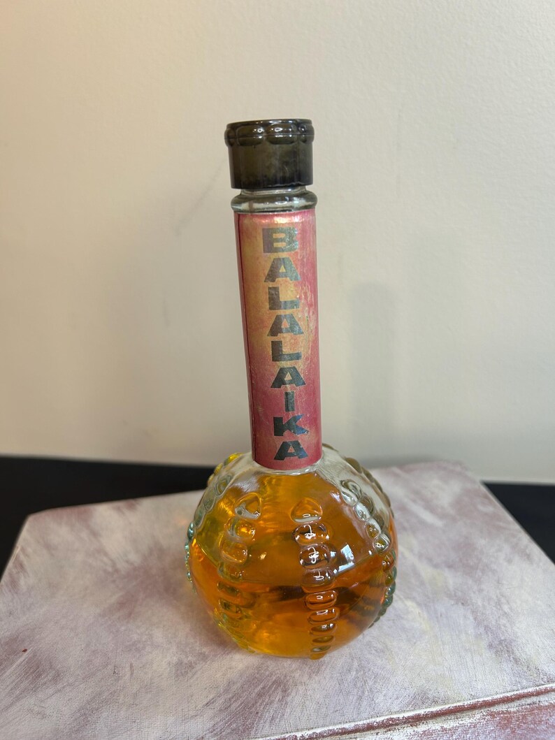 May include: A clear glass bottle with a textured, round base and a long, thin neck. The bottle contains a golden liquid and has a black cap. The bottle has a red label with the word "BALALAIKA" printed in gold letters.