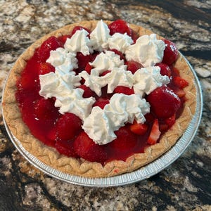 Puede incluir: Una tarta de fresas recién horneada en una lata plateada. La tarta presenta una corteza dorada, un relleno de fresas rojo vibrante y montones de crema batida blanca. La tarta está sobre una superficie moteada marrón y negra.