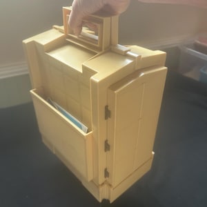 May include: A vintage, mustard-yellow briefcase with a rectangular shape and a handle. The briefcase has a front pocket and a hinged door on the side. The handle is held by a person.