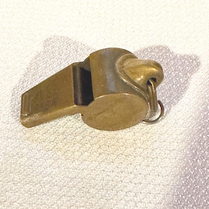May include: A vintage brass whistle with a ring for attachment. The whistle has a rectangular mouthpiece and a rounded body. The text "ACME MADE IN ENGLAND" is visible on the mouthpiece. The whistle is a warm, antique gold color.