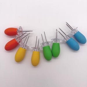 May include: A set of eight corn cob holders with rounded, ergonomic handles in orange, yellow, green, and blue. Each holder features two sharp, parallel stainless steel prongs attached to a clear plastic base, designed for securing corn on the cob.