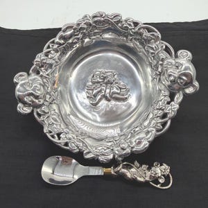 May include: A silver-toned metal bowl featuring a relief of two teddy bears in the center and bear heads on the sides, surrounded by a decorative vine border. A matching silver-toned spoon with a bear-shaped handle is positioned below the bowl.