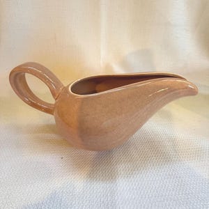 May include: A peach-colored ceramic gravy boat with a curved spout and a looped handle. The gravy boat has a speckled finish and a glossy sheen. The handle is rounded and easy to grip. The boat is designed for serving sauces and gravies.
