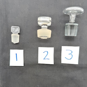 May include: Three clear glass bottle stoppers with different designs. The stoppers are numbered 1, 2, and 3 on white cards. The stoppers have a variety of shapes, including a small, rectangular stopper, a stopper with a rounded top, and a stopper with a flared base.