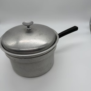 Puede incluir: Una cacerola vintage de aluminio con tapa y asa negra. La olla es de color plateado claro y tiene forma redonda. La tapa tiene un pequeño pomo redondo. El asa está sujeta al lateral de la olla.