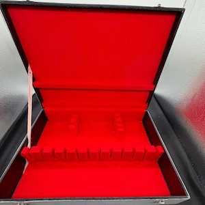 May include: A rectangular storage case with a black exterior and a vibrant red interior lining. The case features a hinged lid, a light-colored fabric support strap, and a series of molded red dividers designed to hold flatware or utensils.