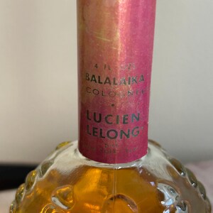 May include: A 4-ounce bottle of Balalaika Cologne by Lucien Lelong. The bottle is clear glass with a textured surface and a pink cap. The label is pink with black text.