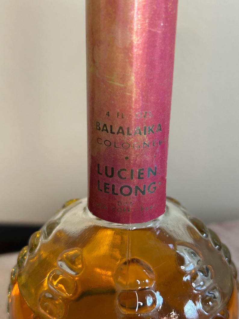 May include: A 4-ounce bottle of Balalaika Cologne by Lucien Lelong. The bottle is clear glass with a textured surface and a pink cap. The label is pink with black text.