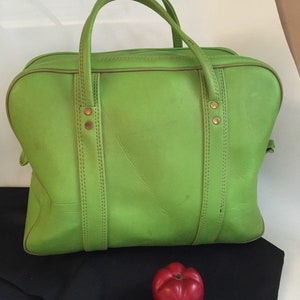 May include: A bright green leather handbag with two top handles and gold accents. The bag has a rectangular shape with rounded corners and is sitting on a black surface.