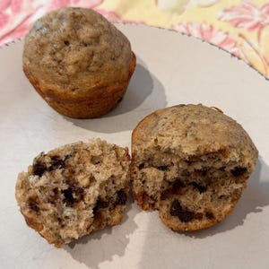 May include: Two freshly baked muffins, one whole and one cut in half, sit on a white plate. The muffins are golden brown with visible chocolate chips. The cut muffin reveals a moist interior with chocolate chips throughout. The plate is on a yellow floral patterned surface.