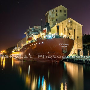 May include: A red cargo ship named "FRONTENAC" docked at a grain elevator at night. The ship is illuminated by lights, and the water reflects the lights. The ship's name and "MONTREAL" are written on the bow.