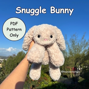 May include: A soft, beige bunny toy with long ears and a pink nose. The text "Snuggle Bunny" is above the bunny, and "PDF Pattern Only" is in a circle to the left. The bunny is a cuddly toy.
