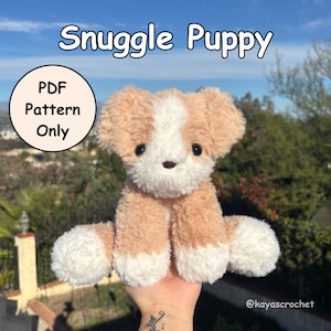 Puede incluir: Un juguete Snuggle Puppy de felpa hecho a mano en tonos beige y blanco. El cachorro tiene ojos grandes y negros y una expresión dulce. La imagen incluye el texto "Snuggle Puppy" y "PDF Pattern Only". El juguete se sostiene frente a un fondo borroso al aire libre.