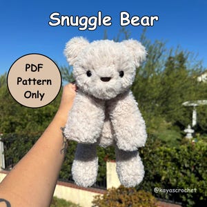 May include: A soft, light grey teddy bear with black eyes and a small, dark nose. The bear is labelled "Snuggle Bear" in white text. A circular graphic reads "PDF Pattern Only." The bear is held up against a backdrop of green foliage and a blue sky.