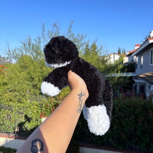 May include: A black and white plush cat toy with fluffy fur. The toy has white paws and a white chest. The background features green foliage and a clear blue sky.