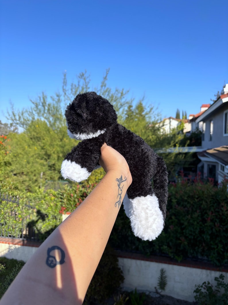 May include: A black and white plush cat toy with fluffy fur. The toy has white paws and a white chest. The background features green foliage and a clear blue sky.