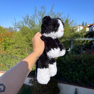 May include: A plush black and white cat toy with yellow eyes and a pink nose. The stuffed animal is held up against a backdrop of green foliage and a clear blue sky.