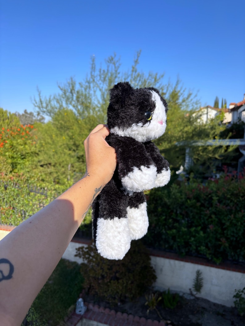 May include: A plush black and white cat toy with yellow eyes and a pink nose. The stuffed animal is held up against a backdrop of green foliage and a clear blue sky.