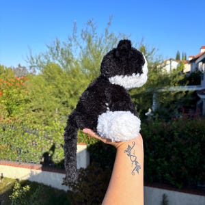 May include: A plush black and white cat toy with a fluffy texture. The cat has a black body, white belly, and white face. The toy is held up against a backdrop of green foliage and a clear blue sky.