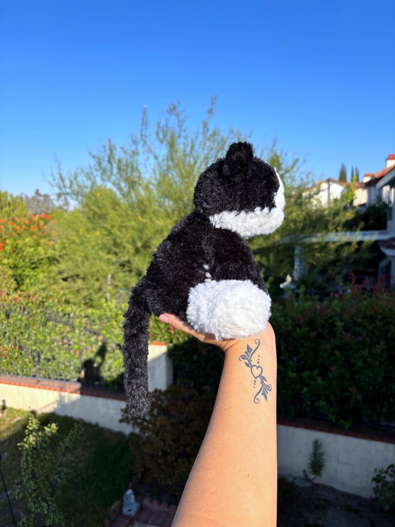 May include: A plush black and white cat toy with a fluffy texture. The cat has a black body, white belly, and white face. The toy is held up against a backdrop of green foliage and a clear blue sky.