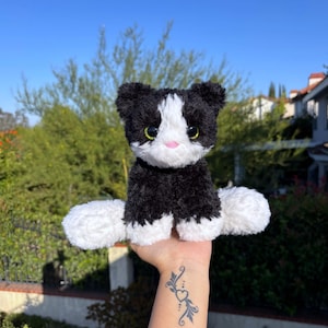 May include: A plush black and white cat toy with a pink nose and green eyes. The stuffed animal has a fluffy texture and is sitting upright. The background features a clear blue sky and green foliage.