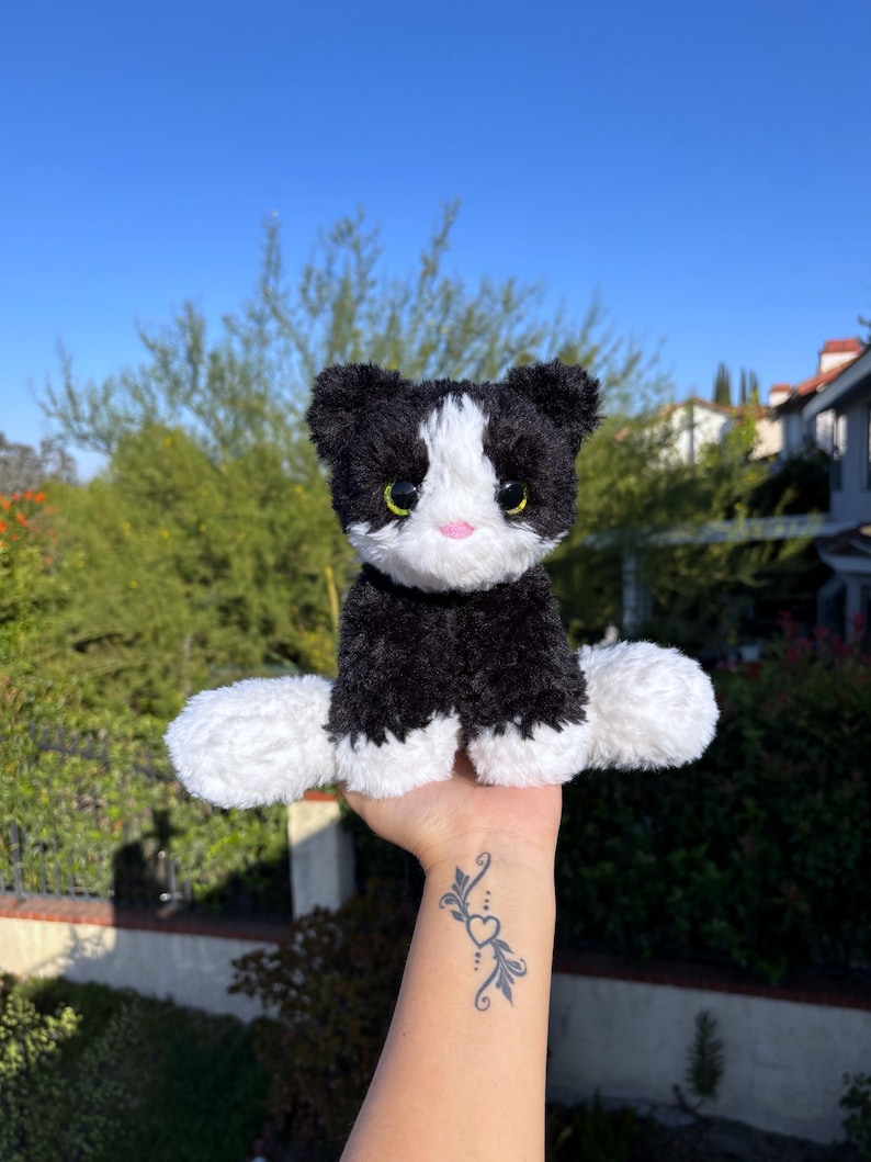 May include: A plush black and white cat toy with a pink nose and green eyes. The stuffed animal has a fluffy texture and is sitting upright. The background features a clear blue sky and green foliage.