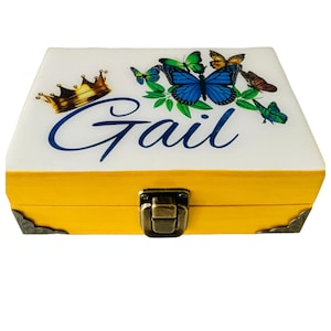 May include: A rectangular yellow wooden box with a white lid featuring the name Gail in blue script, a gold crown graphic, and several colorful butterflies. The box includes a metallic bronze-toned latch and decorative corner accents.