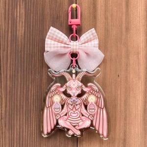 May include: Pink acrylic keychain featuring a seated goat-like figure with wings, holding cupcakes. The keychain has a pink bow and a pink clasp. The background is a wooden surface.